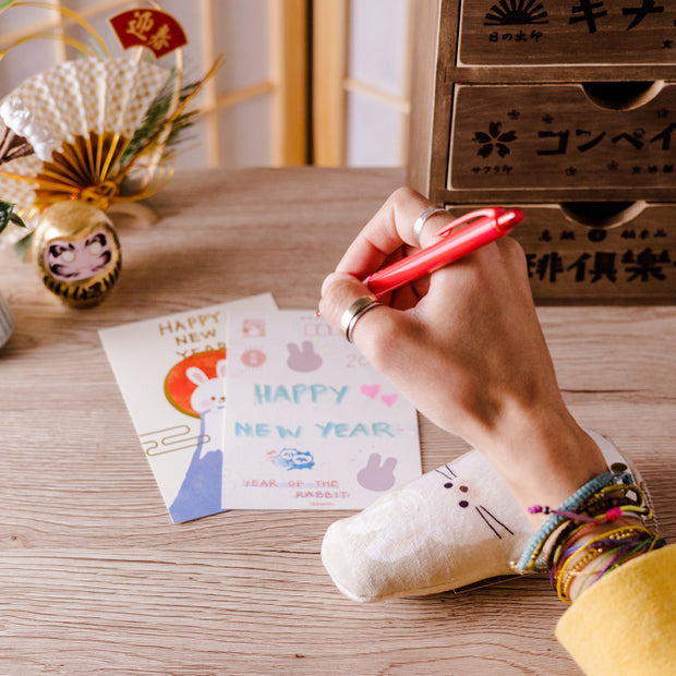 A person uses a red pen from the Inku Stationery Crate by Japan Crate to write a New Year's card at a wooden desk adorned with Japanese ornaments, postcards, and kawaii stationery.