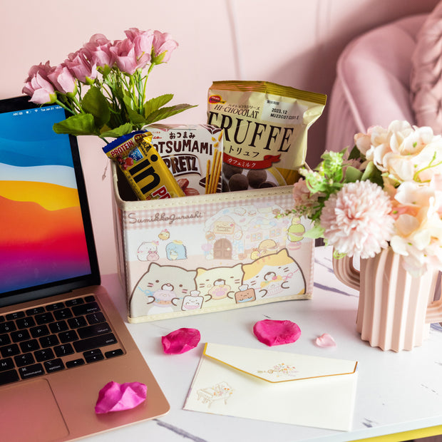 On a white surface sits a desk with a laptop, flowers in vases, an Inku Stationery Crate by Japan Crate filled with kawaii pastel stationery and snacks, and an envelope scattered with rose petals.