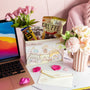 On a white surface sits a desk with a laptop, flowers in vases, an Inku Stationery Crate by Japan Crate filled with kawaii pastel stationery and snacks, and an envelope scattered with rose petals. thumbnail 4