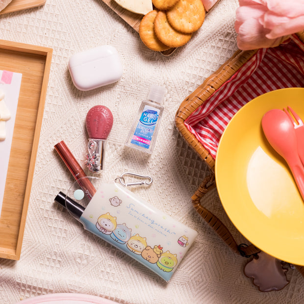 A close-up of picnic items, featuring Japan Crate's Inku Stationery Crate—a kawaii pouch with cartoon designs—plus a yellow plate with pink utensils, crackers, cosmetics, and hand gel on a blanket.