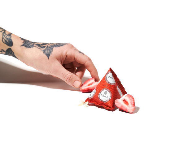 A tattooed hand holds Bokksu White Strawberry 1 Piece alongside sliced chocolate-infused strawberries and a red, pyramid-shaped package on a white background, evoking the distinctive snacks from a Bokksu Snack Box inspired by Nagano, Japan.