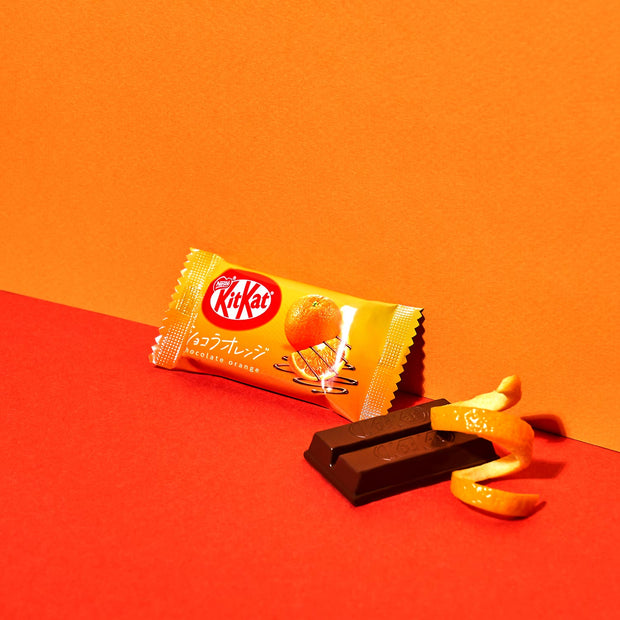 An orange wrapper for Nestle Japan's Japanese Kit Kat: Chocolate Orange (2-Pack) displays a peeled Japanese citrus slice next to two bars on a two-tone background.