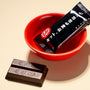 A red bowl with a wrapped Nestle Japan Kit Kat bar labeled in Japanese, beside an unwrapped Nestle Japan Kit Kat: Dark Chocolate on a light beige surface in recyclable packaging. thumbnail 2
