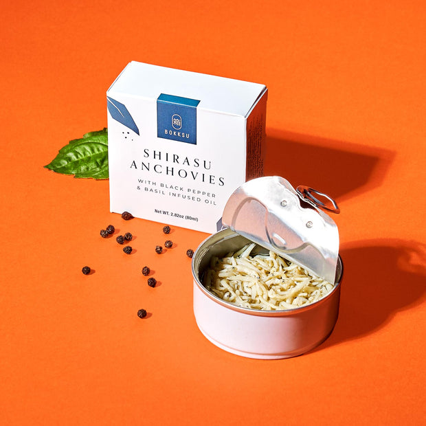 An open can of Bokksu Tinned Shirasu Anchovies with Black Pepper and Basil-Infused Oil rests beside a branded box on an orange surface, accompanied by a basil leaf and peppercorns, suggesting the delightful flavors within.