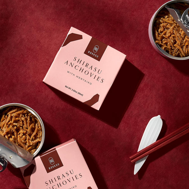 Two open cans of Bokksu Tinned Shirasu Anchovies with Cod Roe (4-Pack), featuring the flavorful Japanese delicacy of cod roe, a pair of red chopsticks, a small plastic spoon, and two unopened boxes of the same product by Bokksu are arranged on a maroon background.