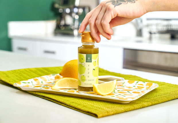 A hand reaches for a bottle of Bokksu Yuzu Hot Sauce by Bokksu, made with fermented yuzu peel, set on a tray with lemon wedges atop a green placemat in a bright kitchen.