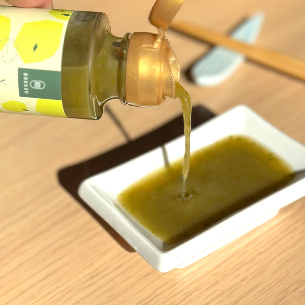 A bottle from the Bokksu Yuzu Hot Sauce (4-Pack) by Bokksu pours its greenish-yellow yuzu kosho sauce into a small rectangular white dish on a wooden surface.
