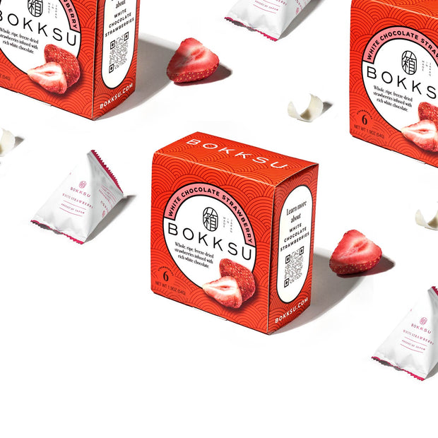 A box of Bokksu White Strawberry 6-Piece Box featuring white chocolate strawberries surrounded by individually wrapped pieces and fresh strawberry halves on a white background.