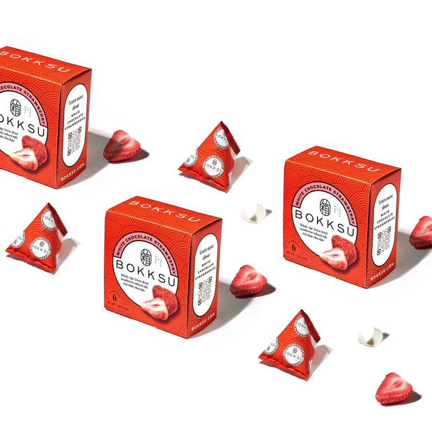 Three red boxes labeled "Bokksu" are displayed with pyramid-shaped chocolates and strawberry slices scattered around them. The Bokksu White Strawberry 6-Piece Box offers delightful Japanese snacks, including strawberry-infused chocolate perfect for indulging your sweet tooth.