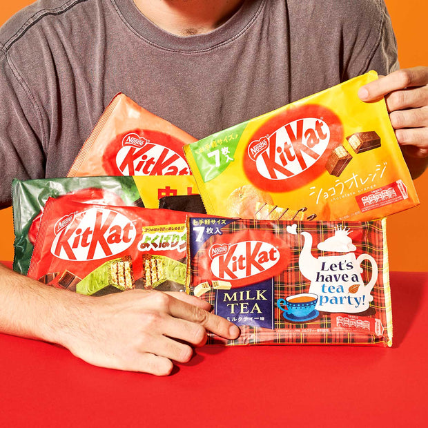 A person holding the Nestle Japan Japanese Kit Kat 6 Flavor Variety Pack (6-pack) including milk tea and other assortments, against a red background.
