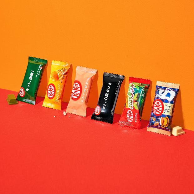 Assorted Japanese Kit Kat 6 Flavor Variety Packs lined up on a contrasting red and orange background from Nestle Japan.
