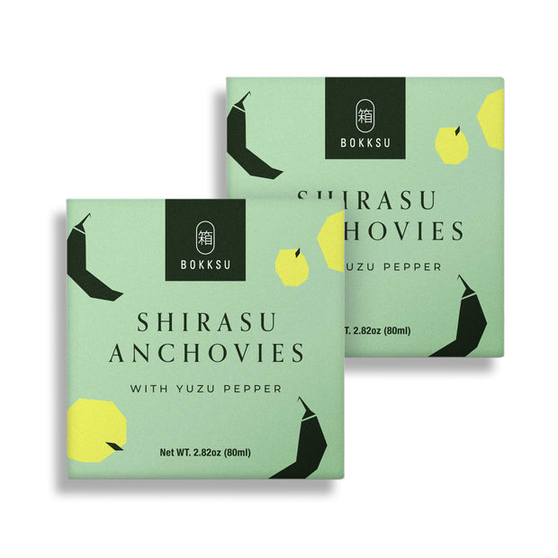 Two green boxes of Bokksu Tinned Shirasu Anchovies with Yuzu Pepper (2-Pack), each with a net weight of 2.82 oz (80 ml). Packed high in protein, the packaging features illustrations of yellow and black geometric shapes.