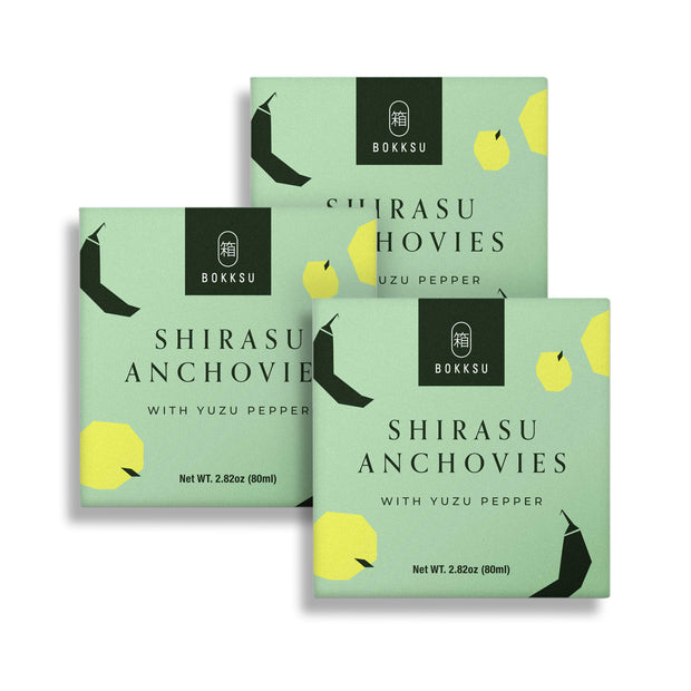 Three boxes of Bokksu Tinned Shirasu Anchovies with Yuzu Pepper (3-Pack), each weighing 2.82 ounces (80 grams), against a white background. The packaging is green with black and yellow text and geometric designs. These snacks are high in protein, making them a nutritious choice.