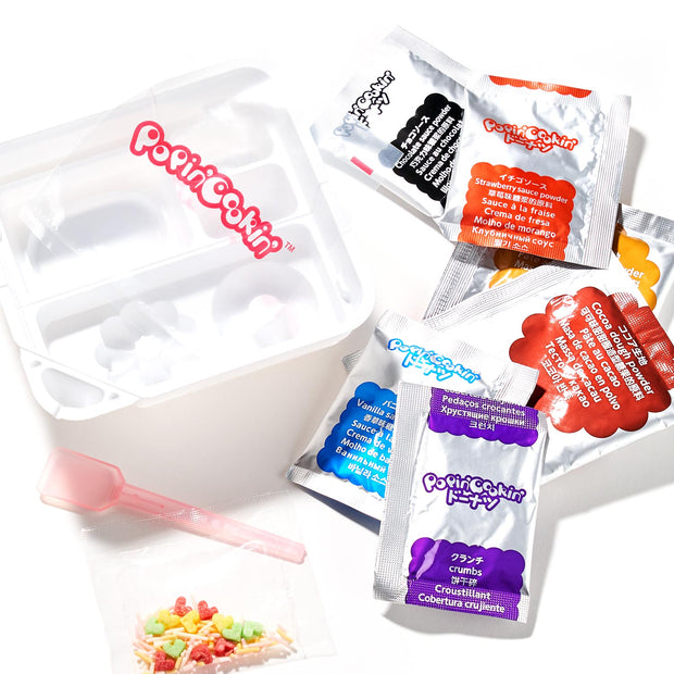 An open Kracie Popin Cookin DIY Candy: Donut kit, specifically a Kracie donut candy kit, with colorful candy packets, a white plastic tray, a small pink spoon, and mixed candy pieces on a white surface.