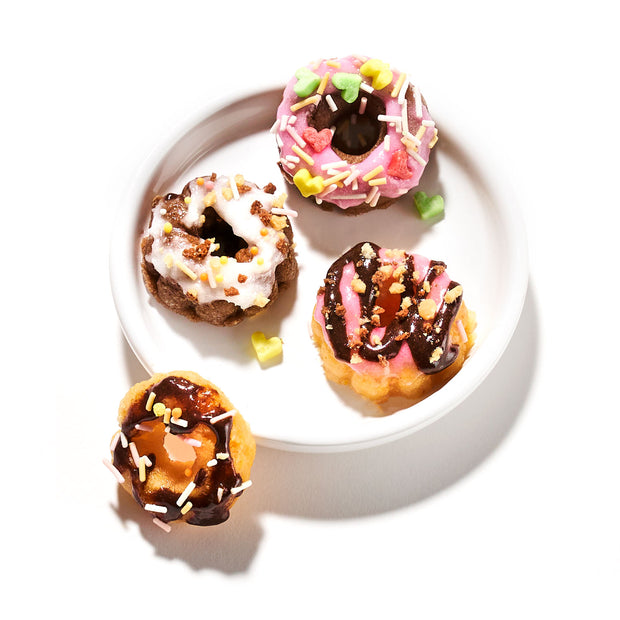 A white plate with four decorated mini doughnuts, crafted from a Kracie Popin Cookin DIY Candy: Donut kit, featuring toppings like chocolate, pink icing, white icing, and colorful sprinkles.