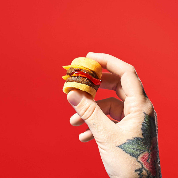 A tattooed hand holds a miniature cheeseburger, reminiscent of the Bokksu Market DIY Candy Kit Pack (6-Pack), set against a bright red background.