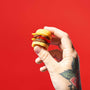 A tattooed hand holds a miniature cheeseburger, reminiscent of the Bokksu Market DIY Candy Kit Pack (6-Pack), set against a bright red background. thumbnail 2