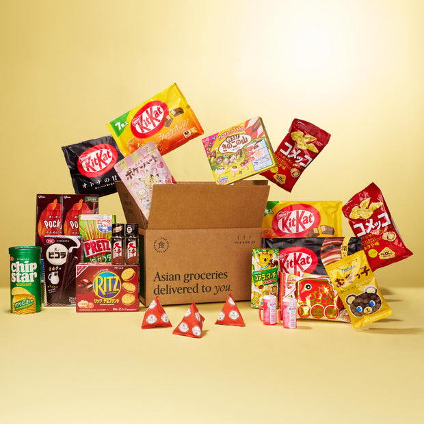 A Bokksu Market Discovery Pack box labeled "Asian groceries delivered to you" sits on a yellow background, surrounded by Asian snacks like KitKat, Pocky, Pretz, Ritz, ramen, chips, candies, and drinks.