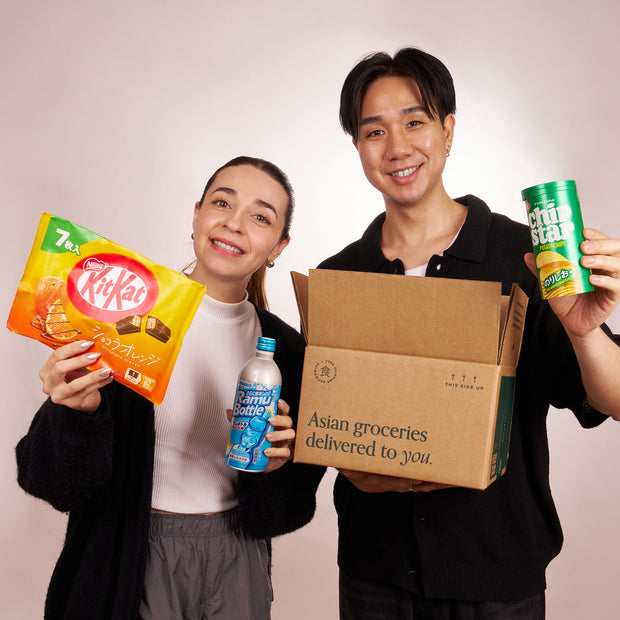 Two people hold the Discovery Pack from Bokksu Market, featuring Asian snacks like instant ramen and mochi, along with a drink and an open "Asian groceries delivered to you" box, all set against a plain background.
