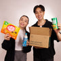 Two people hold the Discovery Pack from Bokksu Market, featuring Asian snacks like instant ramen and mochi, along with a drink and an open "Asian groceries delivered to you" box, all set against a plain background. thumbnail 6