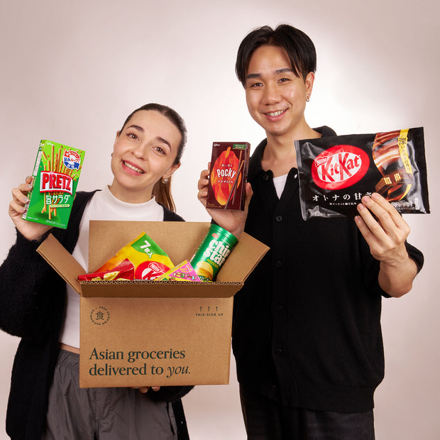 Two people hold Asian snacks, like instant ramen, behind an open Bokksu Market box labeled "Discovery Pack," with packaged treats such as mochi and more Asian groceries visible.