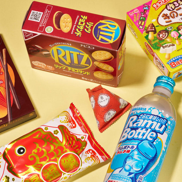 The Bokksu Market Discovery Pack includes Japanese favorites like Ritz crackers, Ramune soda, fish-shaped wafer, Pocky, chocolate candy, and instant ramen—all displayed on a vibrant yellow background.