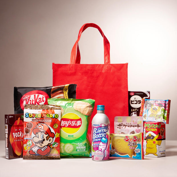 The Snack Attack Lucky Bag from Bokksu Market features a reusable tote behind an assortment of Japanese snacks like KitKat, Lay’s chips, Pocky, Ramune soda, and various colorful candies and cookies.