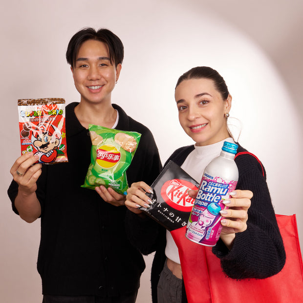 Two people hold up Bokksu Market's Snack Attack Lucky Bag, packed with Japanese snacks and drinks like chips, KitKat, and Ramune soda—perfectly ready to enjoy or carry in a reusable tote—against a plain background.