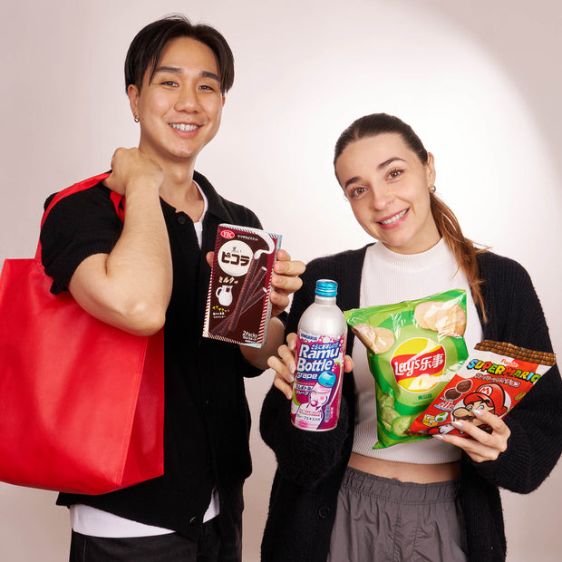 Two people stand side by side, smiling at the camera while holding a Bokksu Market Snack Attack Lucky Bag filled with Japanese snacks and drinks against a plain background.