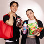 Two people stand side by side, smiling at the camera while holding a Bokksu Market Snack Attack Lucky Bag filled with Japanese snacks and drinks against a plain background. thumbnail 6