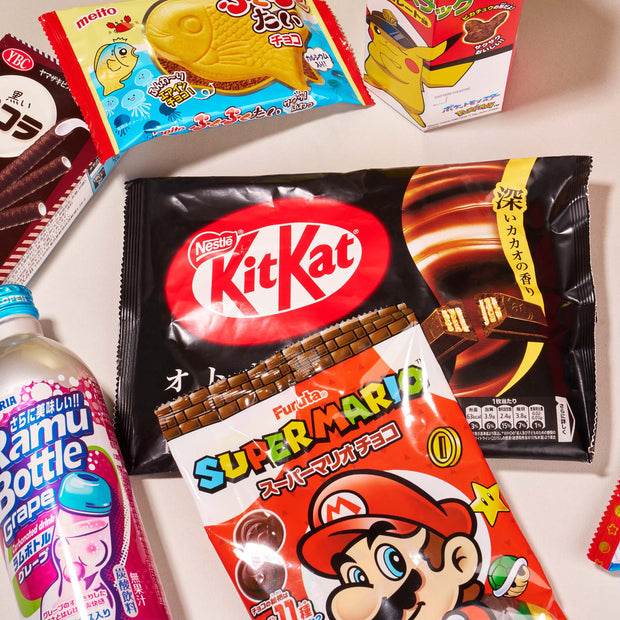 The Snack Attack Lucky Bag by Bokksu Market is filled with Japanese snacks like a black KitKat bag, Super Mario chocolate, Ramune grape soda, and assorted colorful treats displayed on a light surface.