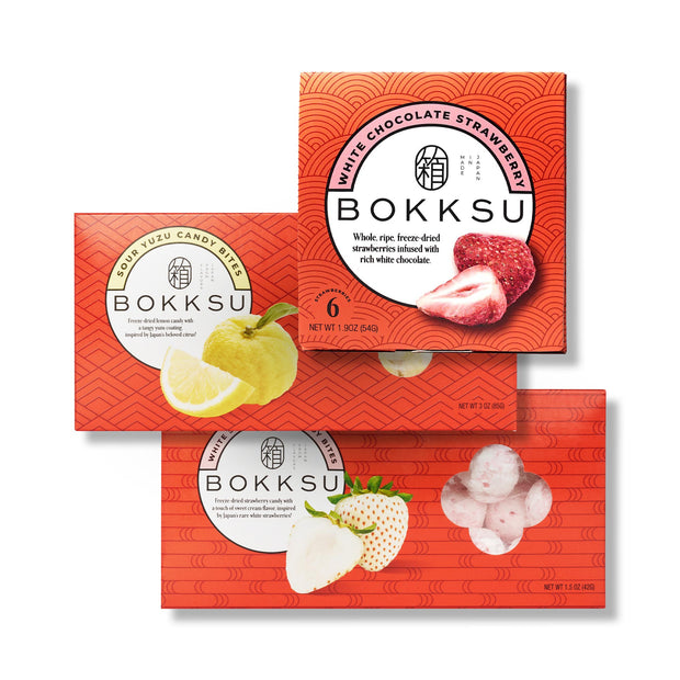 Three Bokksu Market Japanese Candy Bites Trio Delight boxes are stacked, highlighting white strawberry and sour yuzu snacks, with fruit images displayed on the packaging.