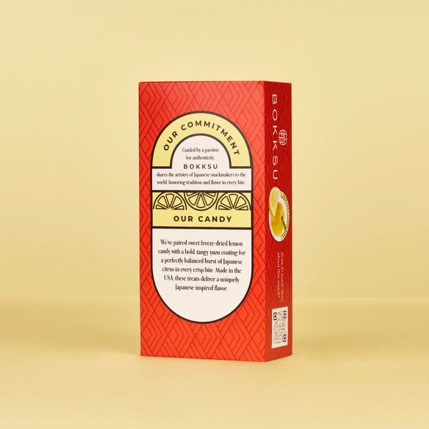A rectangular red Bokksu Market box stands upright against a yellow background, showing product info and a lemon slice image—hinting at Sour Yuzu Japanese Candy Bites (4-Pack) inside.