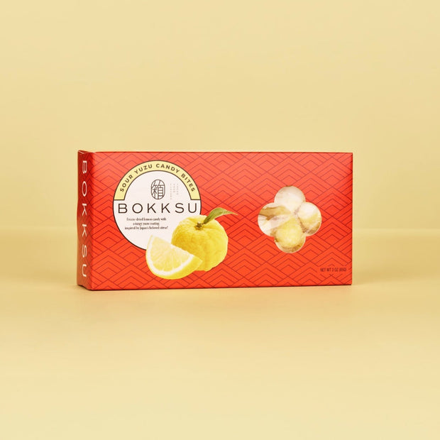 A red box of Bokksu Market's Sour Yuzu Japanese Candy Bites (4-Pack) rests on a plain yellow background, displaying images of Japanese citrus fruit and candies on the front.
