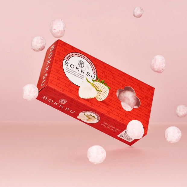 A red Bokksu Market box featuring White Strawberry Japanese Candy Bites sits on a pink background, surrounded by floating freeze-dried treats.