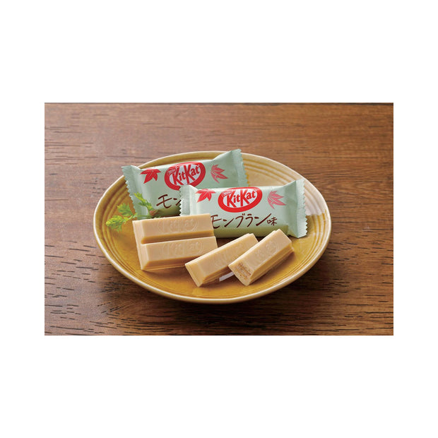 A wooden table holds a plate with two Bokksu Market Japanese Kit Kat: Mont Blanc bars and two wrappers, one open. This seasonal treat features the distinct Mont Blanc flavor.