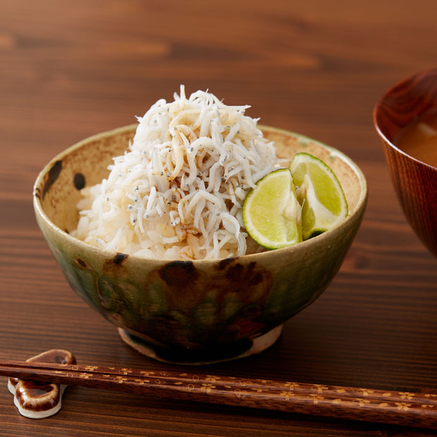 A bowl with rice topped with Bokksu Tinned Shirasu Anchovies with Yuzu Pepper (2-Pack), accompanied by chopsticks and a hint of yuzu kosho on a wooden table.