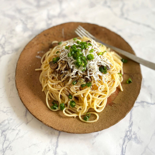 Spaghetti garnished with green peppers, cheese, and Bokksu Tinned Shirasu Anchovies with Cod Roe, served on a brown ceramic plate with a fork.