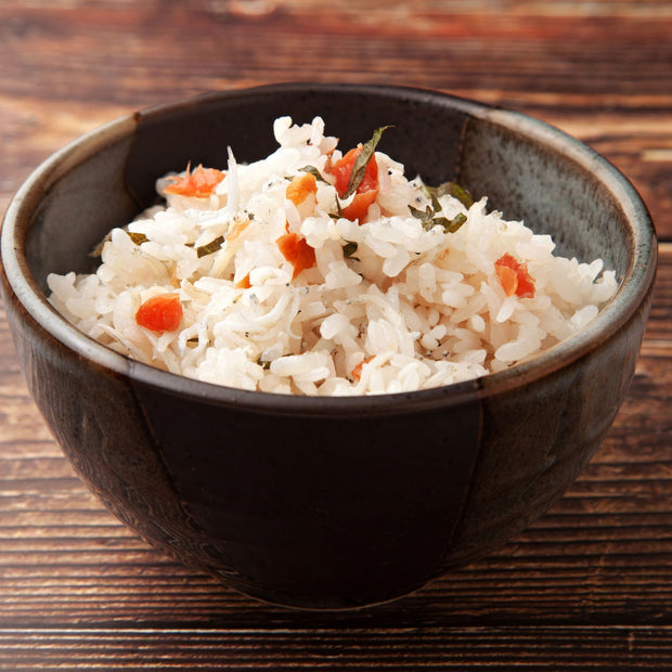 A ceramic bowl filled with cooked white rice mixed with small pieces of vegetables and Bokksu Tinned Shirasu Anchovies with Cod Roe (3-Pack), placed on a wooden surface.