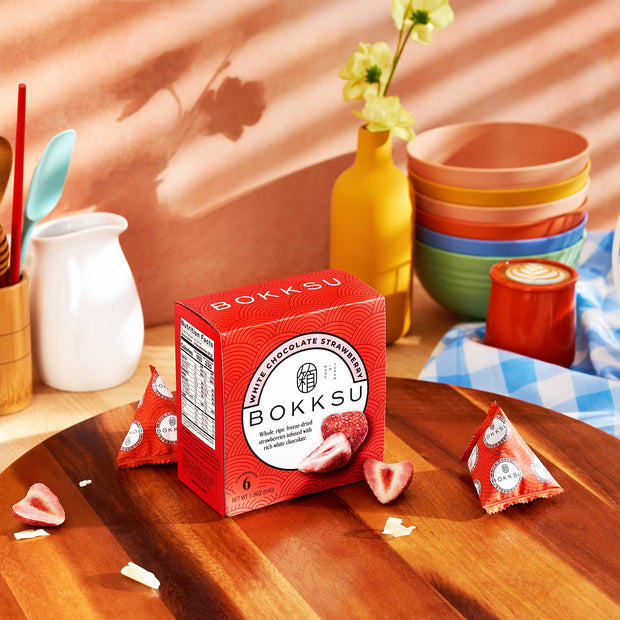 A box of Bokksu White Strawberry 6-Piece treats rests on a wooden table, accompanied by fresh strawberries and kitchenware. These delightful snacks expertly blend strawberry-infused chocolate with an exquisite presentation.