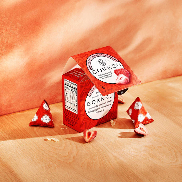 A Bokksu White Strawberry 6-Piece Box is elegantly displayed on a wooden surface, accompanied by individual snack wrappers. Each piece exemplifies the artistry of Japanese snacks, with strawberry-infused chocolate creating an irresistible taste journey.