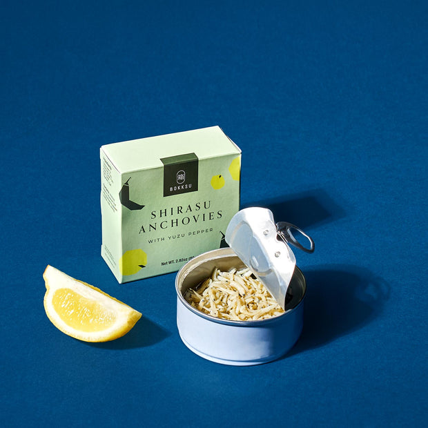 A tin of Bokksu Tinned Shirasu Anchovies with Yuzu Pepper (2-Pack) is open next to its green box packaging and a lemon wedge, arranged on a blue background.