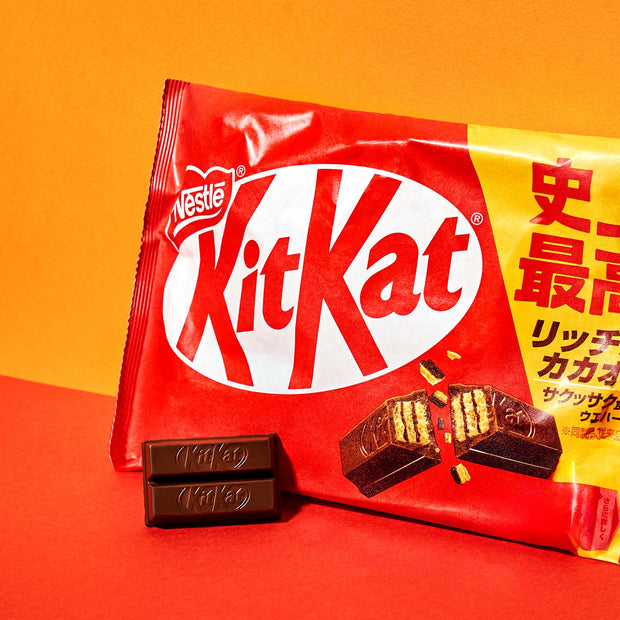A vibrant package of Nestle Japan's Japanese Kit Kat: Original Chocolate (3-Pack) sits on an orange surface, with two milk chocolate pieces attractively displayed in front.
