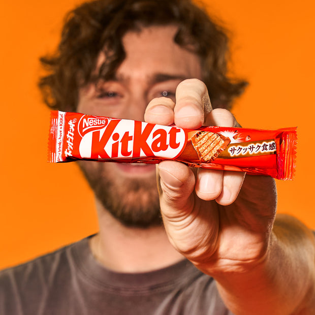 Man holding a Nestle Japan Japanese Kit Kat: Original Bar towards the camera with a blurred smile, against an orange background.