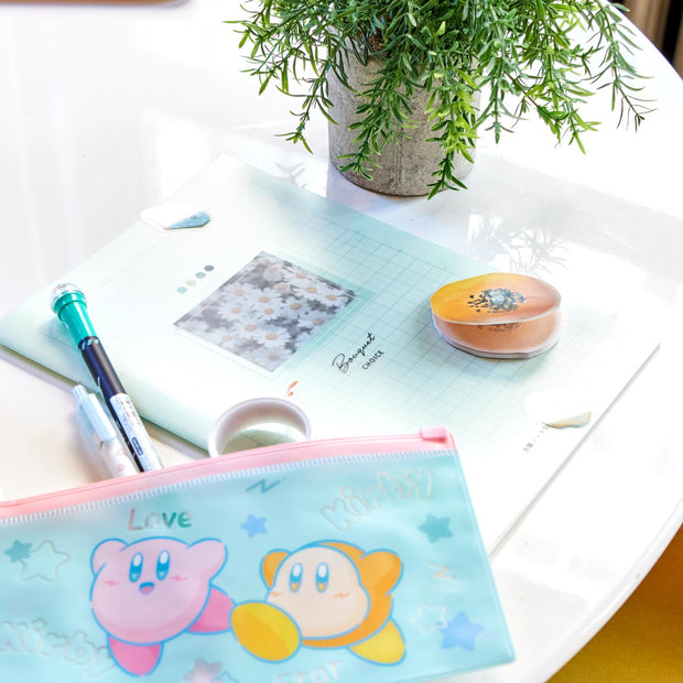 A white table is adorned with the "Kawaii Gift Pack - Stationery Surprises & Japanese Chocolate Treats" by Bokksu, featuring notebooks, pens, pencils, erasers, and a pouch with cartoon characters. A potted plant adds natural charm to the scene.