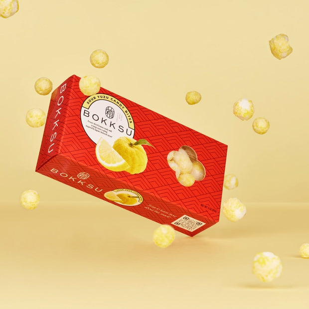 A box of Bokksu Market Sour Yuzu Japanese Candy Bites is displayed with floating yellow candies and a light yellow background. The packaging features images of Japanese citrus yuzu fruit and ginger.