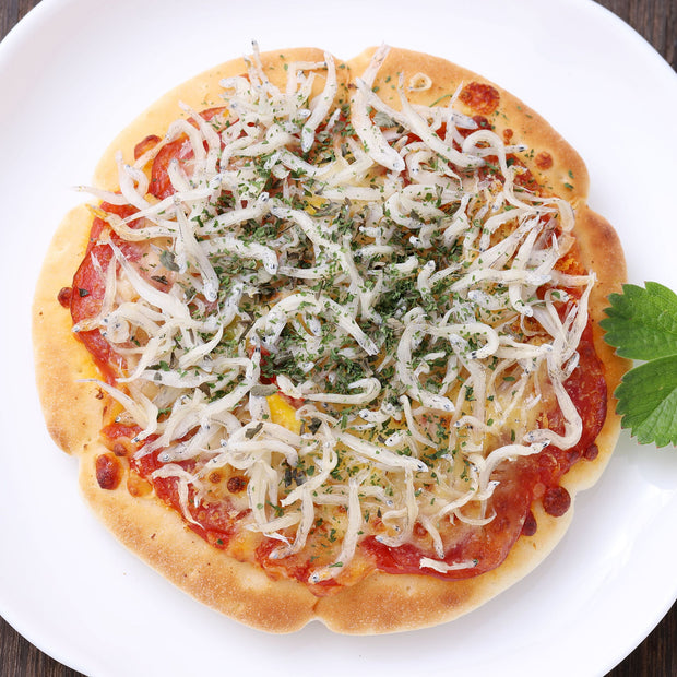 A pizza topped with Bokksu Tinned Shirasu Anchovies with Black Pepper and Basil-Infused Oil (3-Pack), herbs, and a tomato-based sauce, served on a white plate.